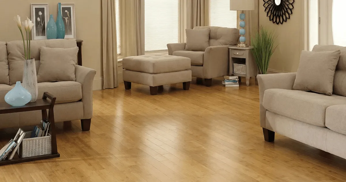 A living room with professionally installed bamboo flooring, beige sofas, a matching ottoman, a side table with a blue lamp, vases, framed pictures, and large windows with blinds and curtains in Overland Park, KS